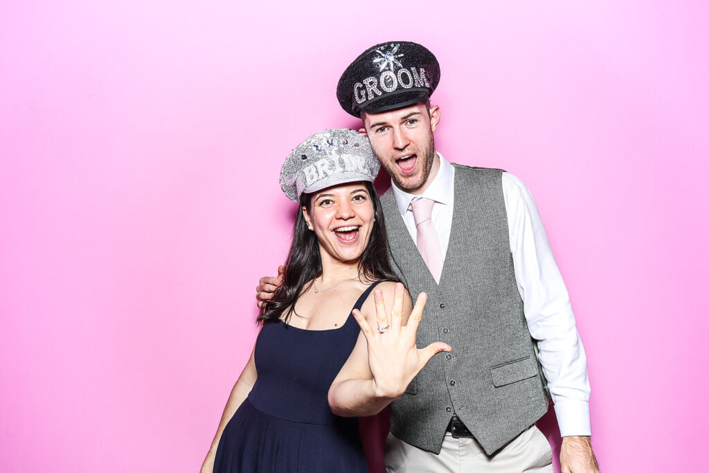 couple posing against a bright bubble pink backdrop for a Wedding Photo Booth with Bride and Groom Hat Props during a wedding party entertainment