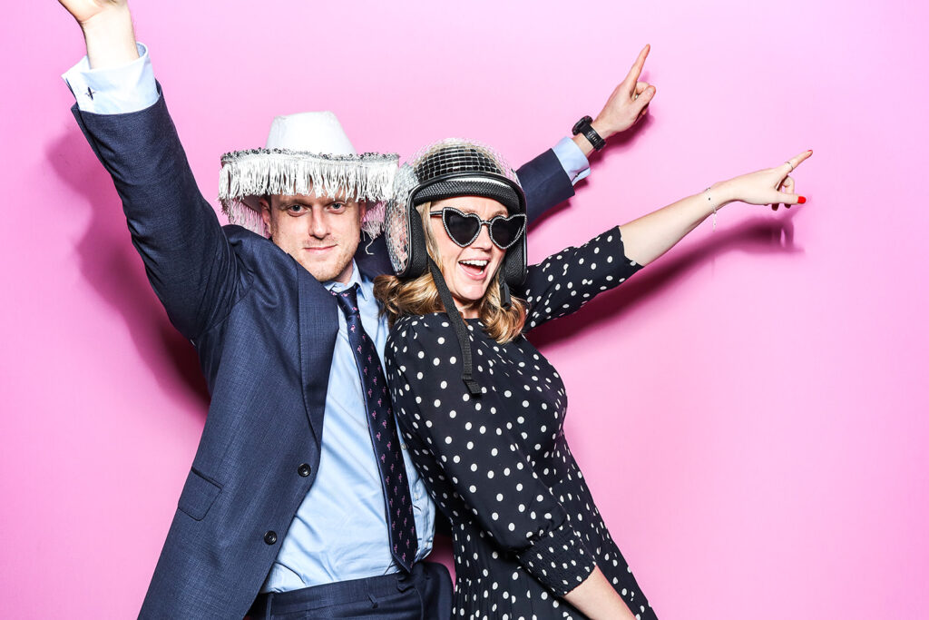 couple posing against a bright bubble pink backdrop for a Wedding Photo Booth with festival Hat Props during a wedding party entertainment