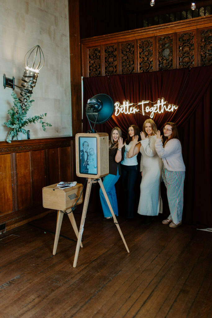 4 ladies posing with their engagement ring, for a luxury wooden photo booth- the icon with a custom rear screen and wooden print box, in front of a burgundy drape backdrop with a "better together" neon sign hire for a wedding event around the cotswolds
