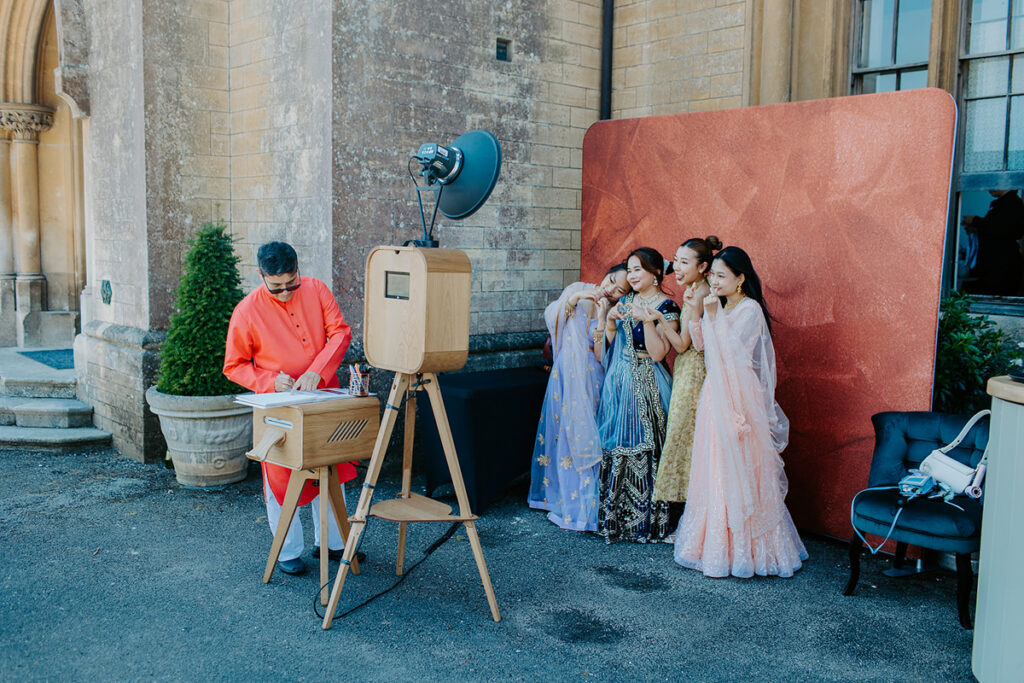 guests posing for a premium wooden photo booth setup with props, and maroon  backdrop for an outdoor garden party at Grittleton House wedding venue in Wiltshire 
