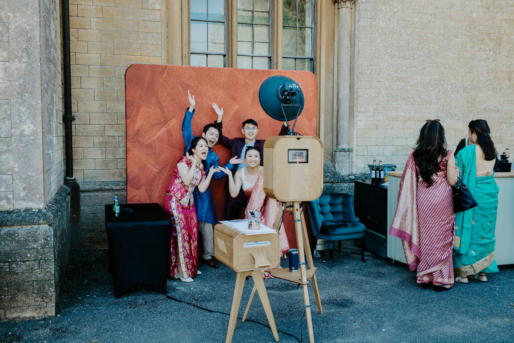 guests posing for a premium wooden photo booth setup with props, and maroon  backdrop for an outdoor garden party at Grittleton House wedding venue in Wiltshire 
